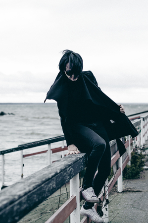fashionable portrait of a young brunette woman in black clothes, jeans T-shirt, coat and sunglasses, in a Gothic style sad mood. on an empty beach, on an autumn cold overcast dayの写真素材