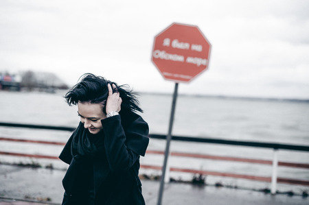 fashionable portrait of a young brunette woman in black clothes, jeans T-shirt, coat and sunglasses, in a Gothic style sad mood. on an empty beach, on an autumn cold overcast dayの写真素材