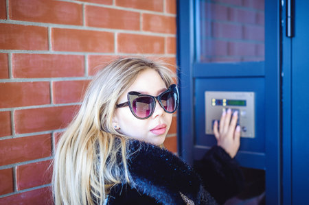 young blond woman in a fur coat, dials the code on the doorphone panel, presses  buttons with her...