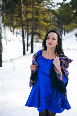 stylish brunette woman with dark hair in a blue dress posing in the open frosty air in a forest among the trees in a traditional Russian wool shawlの写真素材