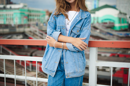 street portrait of a young attractive emotional girl with curly slips dressed in a trendy blue jeans suit on a style walking outdoors against the backdrop of a train station. hand positionの写真素材