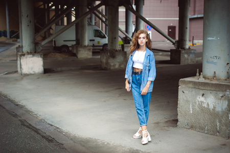 street portrait of a young attractive emotional girl with curly slips dressed in a trendy blue jeans suit on a style walking in the open airの写真素材