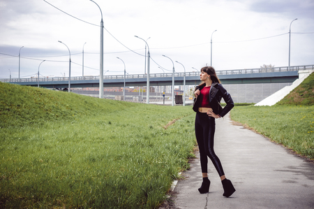 young beautiful stylish woman in fashionable black clothes, a burgundy cardigan and a dark leather jacket posing outdoors near the main road junctionの写真素材