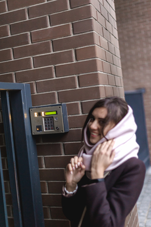 brunette woman calls into the intercom on the street, autumn day in warm clothesの写真素材