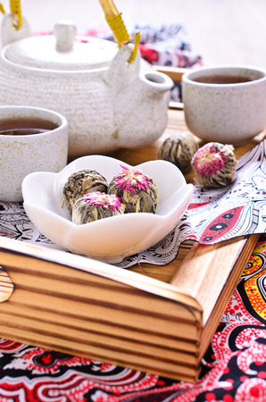 Green tea in the form of a ball with a red flower in a ceramic bowl on a wooden tray surrounded by the kettle and cups made of ceramicの写真素材
