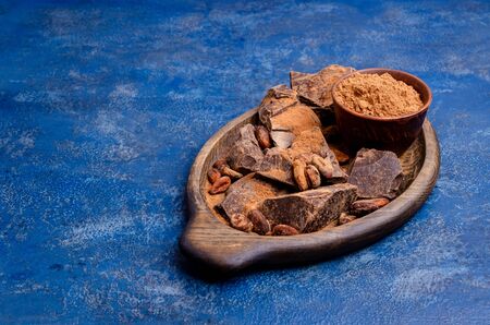 Organic chocolate slices with cocoa beans and brown powder in a wooden dish on a blue background. Selective focus.の写真素材