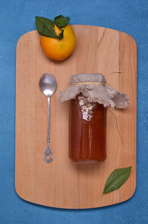 Traditional orange jam in a glass jar on a wooden board on a blue background. Top view.