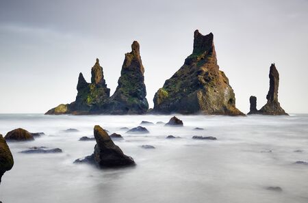 Famous tourist landscape with basalt rock formations Troll toes (Trolls fingers) on black beach. Ocean waves flow around stones. Reynisdrangar, Vik, Iceland, Atlantic Ocean, Europe. Travel postcard.の写真素材