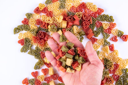 Female hand with handfull of tricolored pasta over heart shaped pasta on white.の写真素材