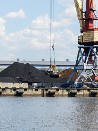 The bucket of a harbor crane in a large pile of black coal at the waters edge