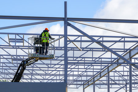 Worker in self propelled articulating boom lift basket in uniform and safety protective equipment on a building steel framework backgroundの写真素材