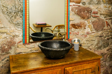 Vintage style bathroom interior with black round ceramic sink with brass faucet on top of old wooden cabinet, stone wall backgroundの写真素材