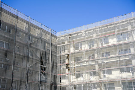 Multi storey apartment building facade renovation and thermal insulation works, scaffolding with gray safety net against a blue sky backgroundの写真素材