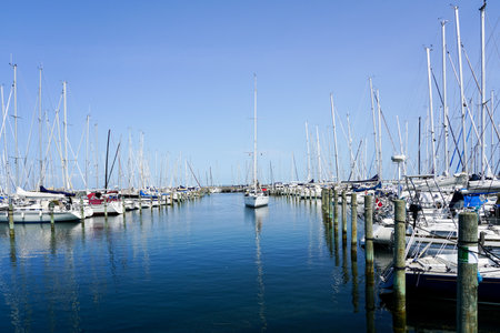 A sailboat floats into the marina channel where yachts are moored on both sides, blue sky backgroundの写真素材