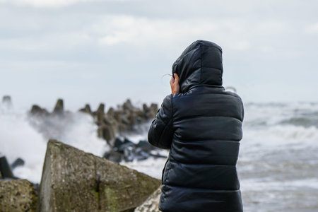 A woman in a black jacket with a hood holds the hood in a strong wind and looks at the stormy sea, rear viewの写真素材