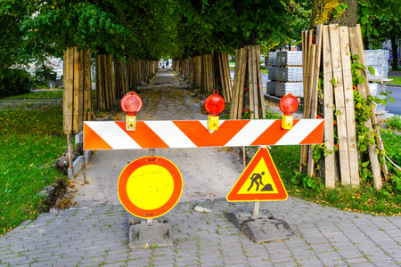 Red and white striped safety barrier with warning signs and red lights, protection of tree trunks from damage with board coversの写真素材