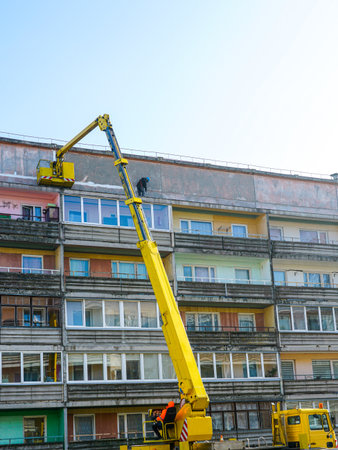 Construction workers use a yellow boom lift to repair and restore the top facade of an old apartment building on a sunny dayの写真素材