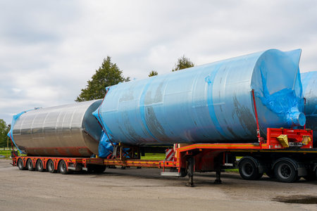 Large stainless steel industrial tanks secured on a flatbed trailer, wrapped in blue plastic for protection during transport and deliveryの写真素材