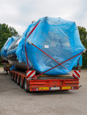 Flatbed truck carrying oversized stainless steel industrial tank wrapped in blue plastic cover and secured with straps for safe transportの写真素材