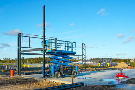 A scissor lift at a construction site with steel frames and building materials. Industrial equipment for construction, engineering, and development projectsの写真素材
