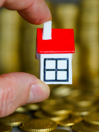 Close-up of a hand holding a miniature house model above stacked gold coins, symbolizing real estate, mortgage, investment, property market, and savingsの写真素材