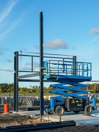 A blue scissor lift positioned at a construction site with a steel frame structure. Industrial equipment for building, engineering, and development projectsの写真素材