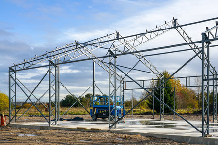 Metal framework of a large industrial warehouse under construction, with scaffolding and blue construction lift on site under bright cloudy skyの写真素材