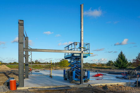 Construction site with steel framework, scissor lift, and scaffolding. Industrial development and modern building process under clear blue skyの写真素材