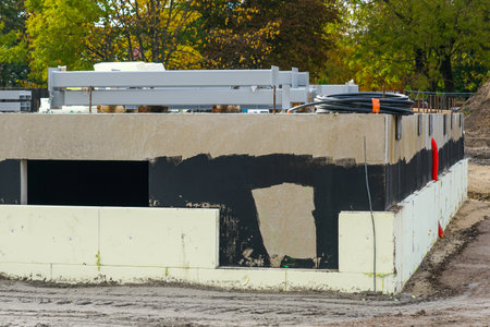 Concrete foundation with insulation panels, black waterproof coating, and red utility pipes installed during building construction phase on site.の写真素材