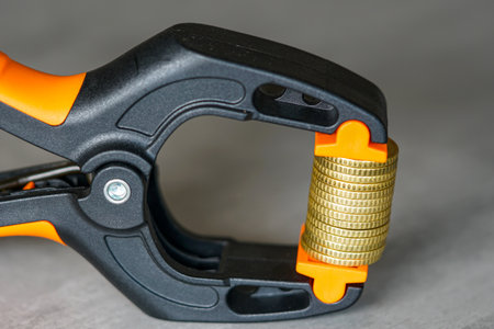 Close-up of gold coins tightly squeezed in a black and orange clamp, symbolizing financial pressure, inflation, economic crisis, and money stressの写真素材