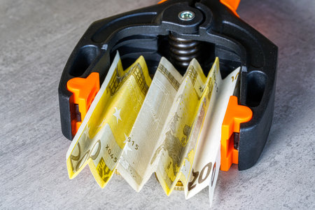 Close-up of euro banknotes tightly squeezed in a black and orange clamp, symbolizing financial pressure, inflation, economy crisis, and cost-of-living stressの写真素材