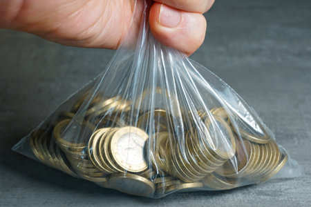 Hand gripping a transparent plastic bag filled with euro coins, representing cash savings, banking, investment, and personal finance conceptsの写真素材