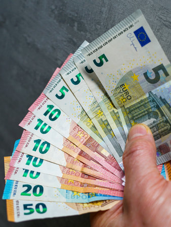 Close-up of a hand holding fan of colorful euro banknotes, symbolizing personal finances, small income, savings, the economy and the European currencyの写真素材