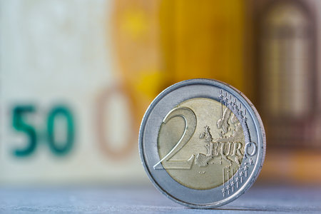 Close-up of a two euro coin standing upright with a blurred fifty euro banknote in the background, symbolizing European currency, finance, and economyの写真素材