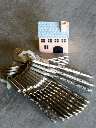 Miniature house figure next to stack of shiny metal keys on a gray surface, symbolizing real estate, property investment, security, and home ownershipの写真素材