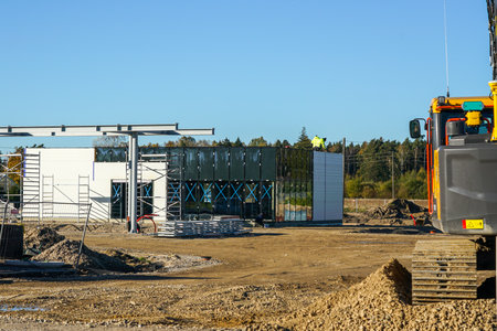 Workers install wall panels at a new fuel station construction site with scaffolding, steel structures, and excavator under a bright blue sky, Latviaの写真素材