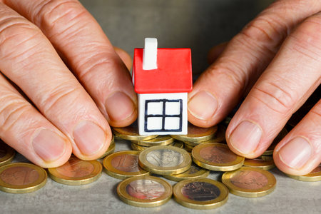 Conceptual image showing hands protecting a small house model on euro coins, representing home savings, real estate investment and financial securityの写真素材