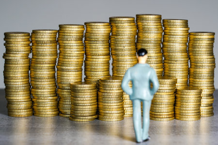 Miniature businessman figure standing before large stacks of gold coins, representing finance, wealth, economic ambition, investment, and prosperityの写真素材
