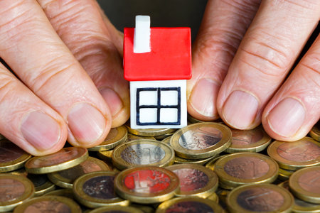 Close-up image showing hands shielding a small house model on euro coins, symbolizing home savings, property investment and financial protectionの写真素材