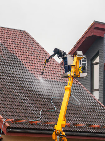 Worker in safety gear cleans a tiled house roof using a high-pressure washer from a hydraulic lift. Roof maintenance and cleaning service in actionの写真素材