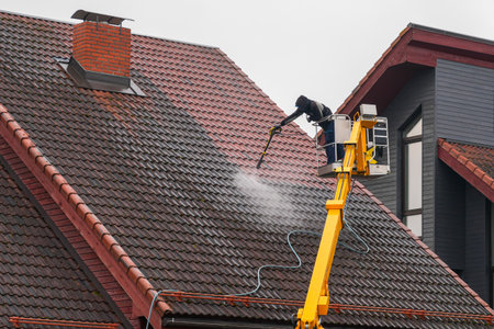 Professional worker cleans a tiled house roof using a high-pressure washer from a yellow lift platform. Roof maintenance and cleaning service conceptの写真素材