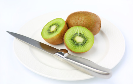 Half of juicy kiwi fruit with knife on white plate against white backgroundの写真素材