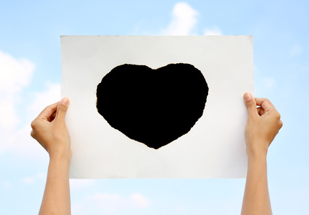 Hands holding paper sheet with Burning in black shape of heart against Cloud sky.の写真素材