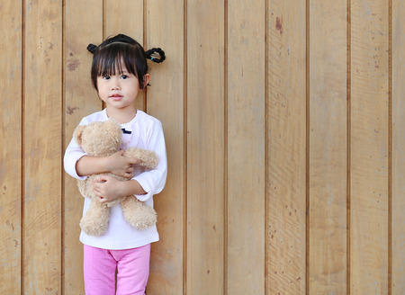 Portrait of cute little girl stand and hugging Teddy Bear against wood plank wallの写真素材