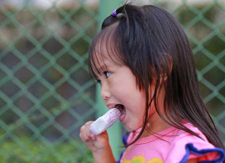 Happy little child girl eating ice-cream in the park against iron mesh backgroundの写真素材