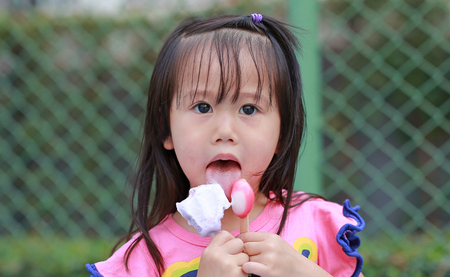 Happy little child girl eating ice-cream in the park against iron mesh backgroundの写真素材