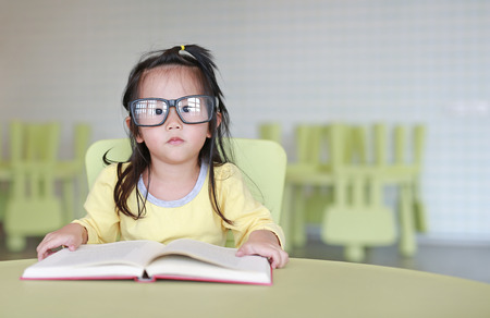 Clever little child girl wearing glasses reading a book in the kids room.の写真素材