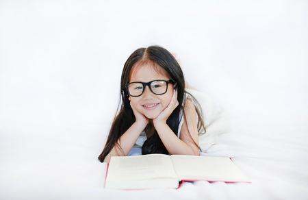 Little Asian kid girl wearing glasses reading hardcover book lying on bed against white backgroundの写真素材
