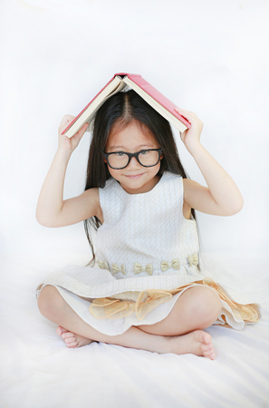 Adorable little Asian girl lying on bed and place hardcover book on her head and looking camera over white backgroundの写真素材