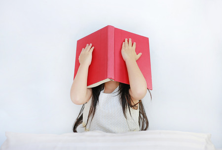 Little Asian child girl hiding face with open hardcover book sitting on bed with pillowの写真素材